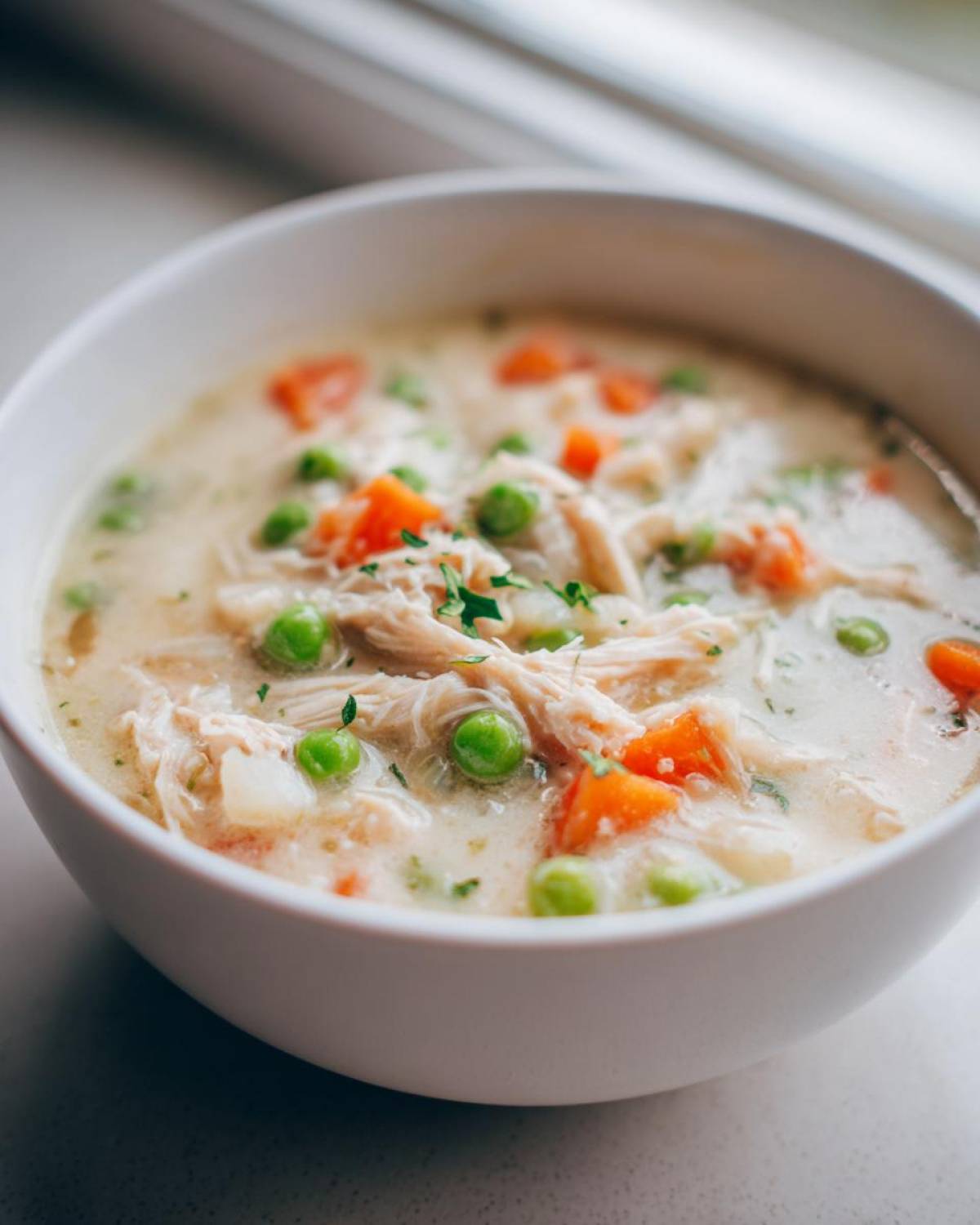 Close-up of a white bowl filled with creamy Slow Cooker Chicken Pot Pie Soup, showing shredded chicken, peas, and carrots.