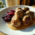 Close-up of five Swedish meatballs smothered in creamy gravy next to a dollop of bright red lingonberry jam.
