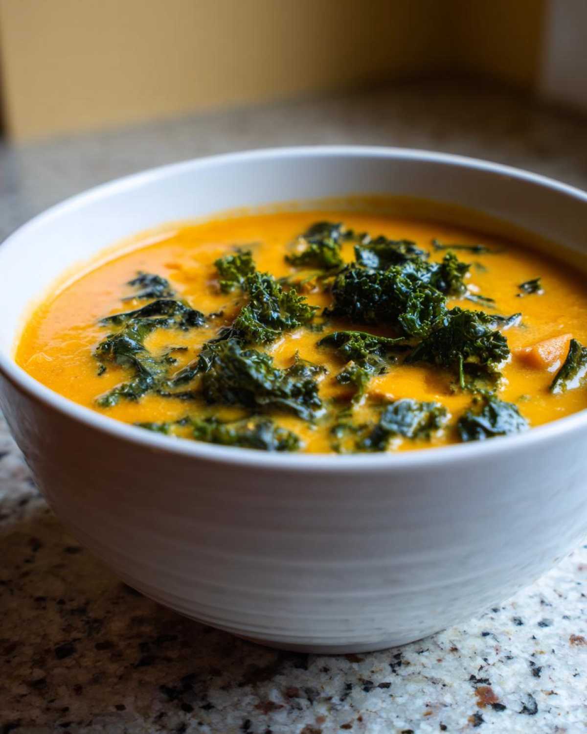 Close-up of a creamy orange Sweet Potato Chowder with Kale floating on top in a white bowl.